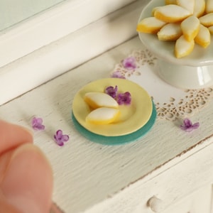 Puede incluir: Una mesa blanca en miniatura con un plato amarillo y azul con galletas de limón en miniatura y flores moradas en la parte superior. Un soporte para pasteles blanco con galletas de limón en miniatura en la parte superior está sobre la mesa.