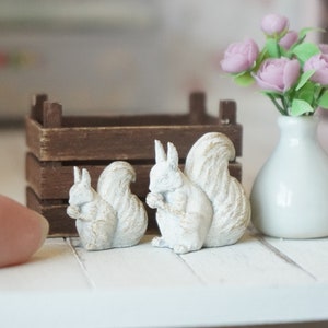 Può includere: Due statuine di scoiattolo in ceramica bianca con finitura usurata, sedute su una superficie di legno bianco. In background, una cassa di legno e un vaso bianco con fiori rosa.