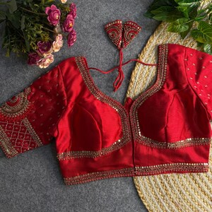 Puede incluir: Una blusa roja con intrincados bordados dorados y lentejuelas. La blusa presenta mangas cortas y un escote en V profundo, con adornos a lo largo de los bordes y las mangas. También se incluye una pieza decorativa a juego.