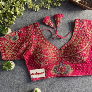 May include: A vibrant fuchsia blouse featuring intricate gold embroidery and peacock motifs. The garment has a deep V-neckline, short sleeves, and a back tie closure with two decorative tassels. A small white tag with the word designer is attached.