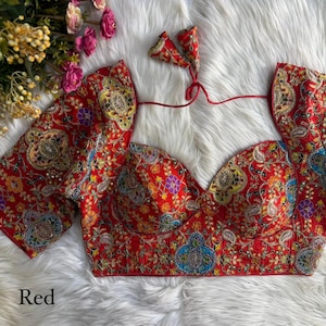 May include: A red, intricately embroidered blouse with short sleeves and a sweetheart neckline. The blouse features detailed floral and paisley patterns in various colors, including gold, blue, and purple. The word "Red" is printed at the bottom.