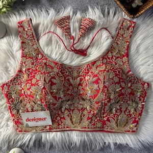 May include: A red sleeveless blouse featuring detailed gold embroidery and sequin accents. The floral pattern adorns the entire front and back. Two decorative pieces, matching the blouse, are connected by a red string. A small tag with the word "designer" is visible.