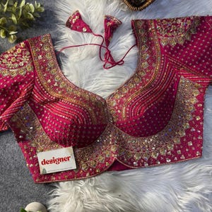 May include: A magenta-colored, heavily embellished blouse with intricate gold embroidery and sequin detailing. The blouse features a deep V-neckline and short sleeves. The word "designer" is printed on a small white tag. The garment is displayed on a white, fluffy surface.