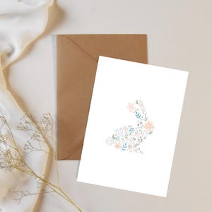 May include: A white greeting card with a floral bunny design in pastel colours, and a brown envelope behind it. The bunny is made up of pink, blue, and white flowers and leaves. The card is on a white surface with a beige fabric and dried flowers.