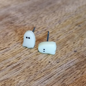 Może przedstawiać: Para białych kolczyków w kształcie ducha z czarnymi oczami. Kolczyki leżą na drewnianej powierzchni.