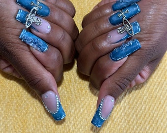 May include: Close-up of hands with long, blue, denim-inspired nails. Each nail is decorated with a silver charm and rhinestones.