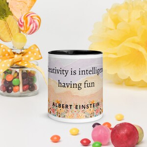 Könnte beinhalten: Ein weißer Keramikbecher mit schwarzem Innenraum und Rand, der einen aquarellartigen Hintergrund in Pastelltönen aufweist. Der Text lautet Creativity is intelligence having fun, gefolgt von ALBERT EINSTEIN über einem bunten Blumenmuster.