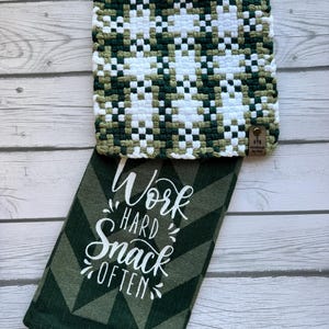 May include: A handmade, square potholder with a woven pattern in white, green, and olive green. A matching green and white kitchen towel with the text "Work Hard Snack Often" is below the potholder.