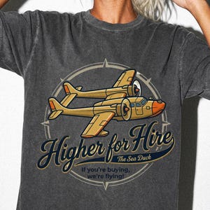 Könnte beinhalten: Ein anthrazitfarbenes Kurzarm-T-Shirt mit einer Grafik eines gelben zweimotorigen Wasserflugzeugs. Der Text lautet Higher for Hire, The Sea Duck und if you're buying, we're flying! Das Design ist vor einem kreisförmigen Kompasshintergrund platziert.