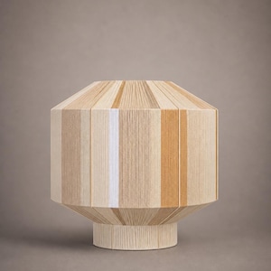 Op de afbeelding: Een decoratieve lamp met een cilindrisch lichaam en een uitlopende voet. De lamp is gemaakt van verticale strengen garen in tinten beige, wit en bruin. De lamp heeft een geometrische vorm en een modern design.