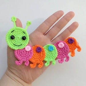 May include: A vibrant, crocheted caterpillar toy. The caterpillar has a green head with black eyes and a smiling mouth, followed by segments in pink, orange, and green. Each segment has small legs and is topped with a contrasting colour.