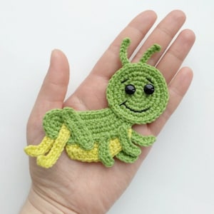 May include: Green crocheted grasshopper with yellow legs and a smiling face. The grasshopper has black eyes and a black stitched smile.