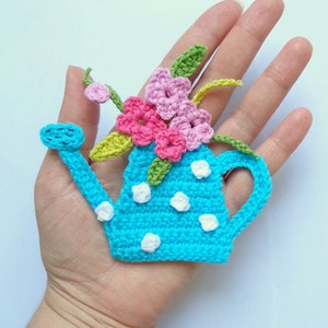May include: A crocheted blue watering can with white polka dots and a bouquet of pink, white, and green flowers.