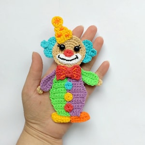 Könnte beinhalten: Eine gehäkelte Clownpuppe mit einem gelben Hut, blauen Haaren, einer roten Schleife und einem grün-violetten Outfit. Die Puppe hat ein lächelndes Gesicht mit schwarzen Augen, einer roten Nase und einem weißen Lächeln.