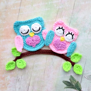 May include: Two crocheted owls with pink and blue hearts on a brown branch with green leaves. The owls have white eyes and black pupils.