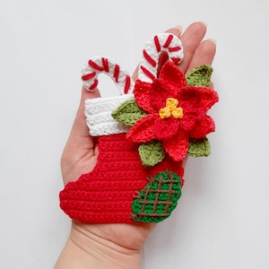 Peut inclure: Une chaussette de Noël rouge en crochet avec un revers blanc, une canne en sucre et une poinsettia rouge avec des feuilles vertes.