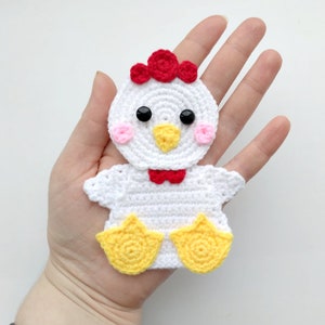 Peut inclure: Poulet crocheté blanc avec des détails rouges et roses, des pieds jaunes et des yeux noirs.