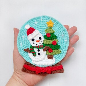Puede incluir: Un adorno de bola de nieve de crochet con un muñeco de nieve y un árbol de Navidad en su interior. La bola de nieve es azul, el muñeco de nieve es blanco con un gorro rojo y una bufanda verde y roja, y el árbol de Navidad es verde con adornos rojos y una estrella amarilla en la cima.
