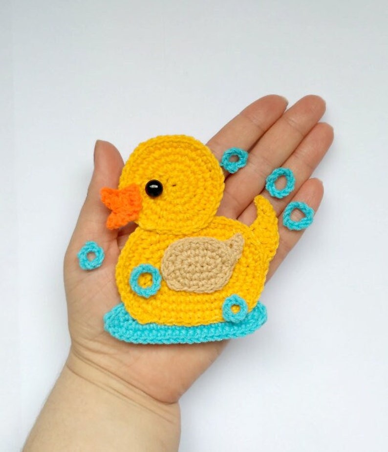 Puede incluir: Un pato de goma amarillo tejido a crochet con una base azul y un pico naranja. El pato est&aacute; rodeado de c&iacute;rculos azules tejidos a crochet, que parecen burbujas.