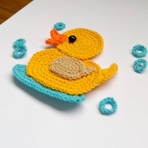 Puede incluir: Un pato de goma amarillo tejido a crochet con un pico naranja y ojos negros, sentado en una tabla de surf azul tejida a crochet. El pato est&aacute; rodeado de anillos azules tejidos a crochet.