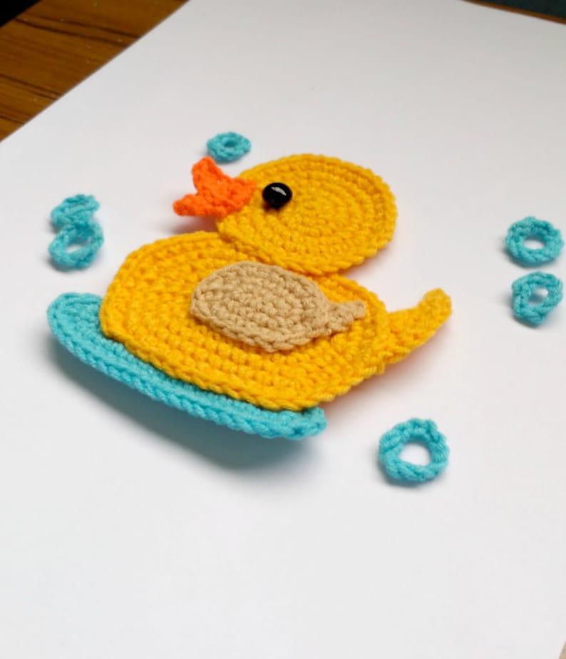 Puede incluir: Un pato de goma amarillo tejido a crochet con un pico naranja y ojos negros, sentado en una tabla de surf azul tejida a crochet. El pato est&aacute; rodeado de anillos azules tejidos a crochet.