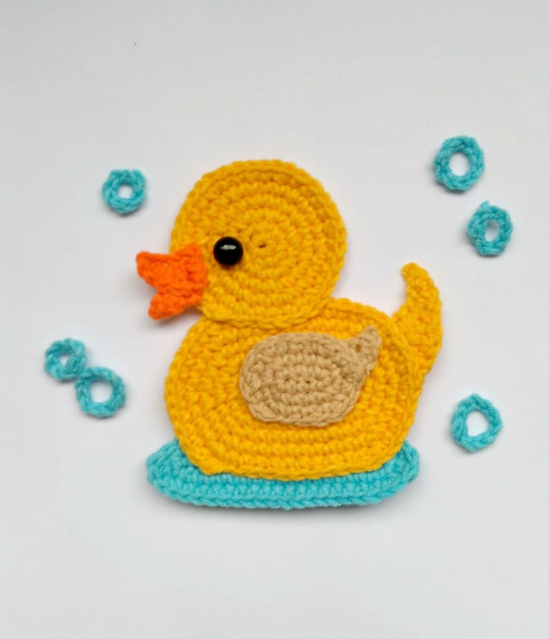 Puede incluir: Un pato de goma amarillo tejido a crochet con la espalda marr&oacute;n y la base azul, rodeado de c&iacute;rculos azules tejidos a crochet.