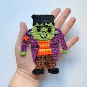 Puede incluir: Un muñeco de Frankenstein de crochet verde con pelo negro, chaqueta morada, camisa a rayas naranja y amarilla, pantalones marrones y zapatos negros.