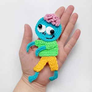 May include: A crocheted blue and green zombie toy with a pink brain, yellow pants, and big white eyes.
