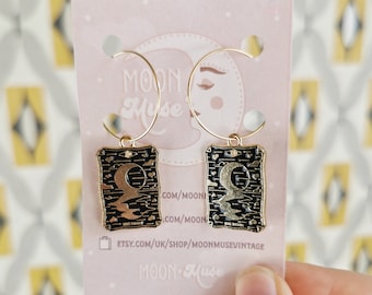 May include: Gold hoop earrings with black and gold enamel charms featuring the The Moon tarot card. The charms have a gold crescent moon and stars on a black background. The card text reads "The Moon". The earrings are displayed on a pink card with the text "Moon Muse" and a website address.