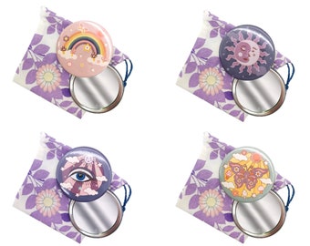 Puede incluir: Cuatro espejos compactos con diferentes diseños. Cada espejo está en una bolsa de cordón floral morado. Los diseños incluyen un arcoíris con nubes, un sol y una luna, un ojo con nubes y una mariposa con nubes.