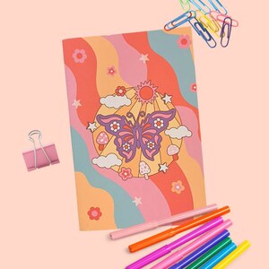 May include: A pink and orange notebook with a retro butterfly design. The notebook is surrounded by colourful paper clips and markers.