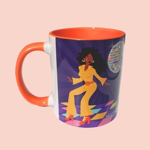 May include: White ceramic mug with an orange handle and interior. The mug features a vibrant illustration of a woman in a yellow jumpsuit dancing in a disco setting, with a disco ball and the word "MOON" visible.