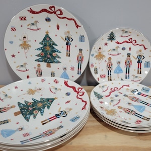 May include: A set of white ceramic plates with a Christmas-themed design. The plates feature illustrations of nutcrackers, ballerinas, Christmas trees, and gift boxes. The plates have a gold rim and a red ribbon border.