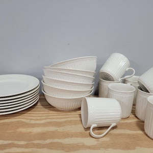 May include: A collection of white ceramic tableware, including plates, bowls, mugs, and small containers. The plates are stacked on the left, while the bowls are stacked in the center. The mugs and containers are arranged on the right.