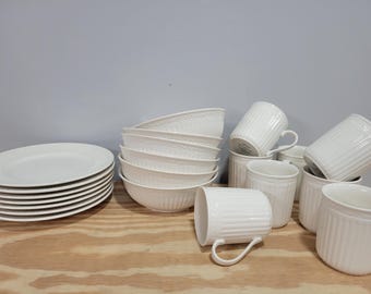 May include: A collection of white ceramic tableware, including plates, bowls, mugs, and small containers. The plates are stacked on the left, while the bowls are stacked in the center. The mugs and containers are arranged on the right.