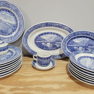 May include: A collection of blue and white ceramic dinnerware featuring landscape scenes. The set includes plates, serving platters, a sugar bowl, a teacup, and saucers. The rims of the plates have a decorative border.