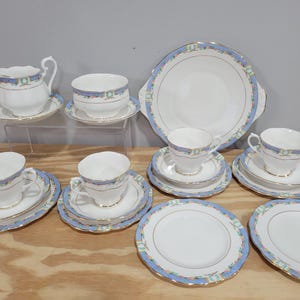 May include: A vintage porcelain tea set with a light blue and gold trim. The set includes teacups, saucers, plates, a creamer, a sugar bowl, and a serving platter. The set is arranged on a wooden surface.