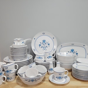 May include: A set of white and blue floral patterned dinnerware, including plates, bowls, cups, and saucers. The set features a blue floral design with a crown motif.