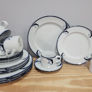 May include: A white porcelain dinnerware set with a navy blue floral design. The set includes plates, cups, saucers, and a sugar bowl. The plates and cups are stacked, and the set is arranged on a wooden surface.