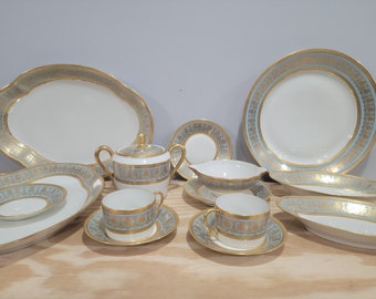 May include: A set of white porcelain dinnerware with gold and light blue trim. The set includes a large oval platter, a smaller oval platter, a round dinner plate, a round salad plate, a teacup and saucer, a creamer, and a sugar bowl.