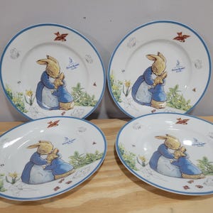 May include: Set of four white ceramic plates with blue trim. Each plate features a colorful illustration of Peter Rabbit and his mother, surrounded by flowers and greenery. The plates have the text "Peter Rabbit" in blue.