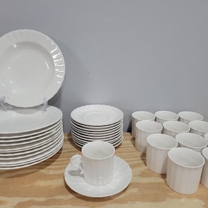 May include: A set of white ceramic dinnerware, including plates, bowls, and cups. The plates are stacked in two piles, and the bowls and cups are arranged in a semi-circle.
