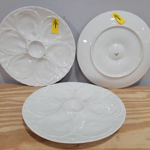 May include: Three white ceramic oyster plates featuring six molded shell-shaped indentations arranged around a central circular well. Two plates show the top surface with shell patterns, while one plate is flipped to show its smooth underside.