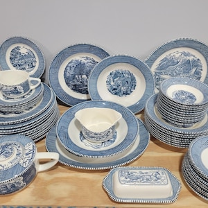 May include: A collection of blue and white ceramic dinnerware featuring landscape scenes. The set includes plates, bowls, a teapot, a creamer, and a butter dish. The plates have a decorative border. The dinnerware is arranged on a wooden surface.