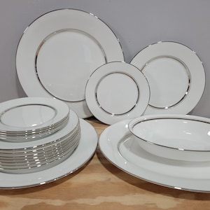 May include: A collection of white ceramic dinnerware with a silver-colored trim. The set includes various sizes of plates, bowls, and serving dishes. The plates are stacked and arranged on a wooden surface.