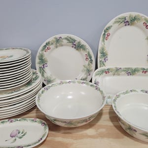 May include: A collection of white ceramic dinnerware featuring a grape and leaf design. The set includes plates, bowls, and serving platters. The plates are stacked in a neat pile, and the bowls and platters are arranged in front. The design includes green leaves and purple grapes.