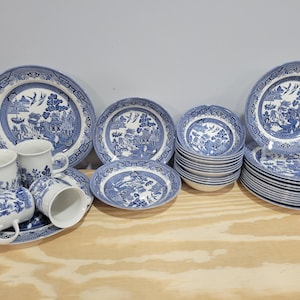 May include: A collection of white ceramic dinnerware featuring a blue willow pattern. The set includes several mugs, large dinner plates, medium-sized bowls, and a stack of smaller bowls, all decorated with intricate blue landscape illustrations.