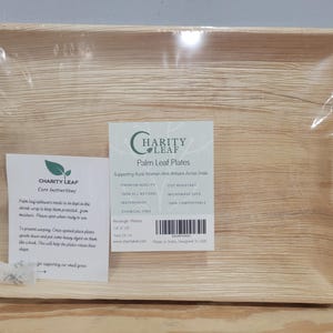 May include: A rectangular palm leaf plate wrapped in clear plastic, featuring a light tan wood-grain texture. A label reads Charity Leaf Palm Leaf Plates, 14 x 10 inches, and a separate care instruction sheet is attached to the front surface.