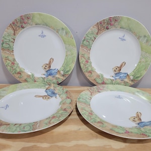 May include: Set of four white ceramic dinner plates with a green and brown border featuring a Peter Rabbit illustration. Each plate has a different Peter Rabbit design.