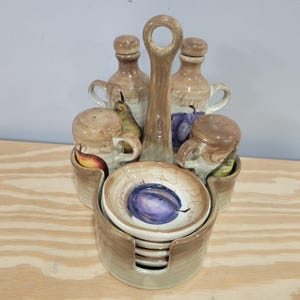 May include: A ceramic kitchen set featuring a handle, two bottles, two salt shakers, and small bowls. The set is painted with fruit illustrations in shades of brown, purple, and green. The set is designed to hold condiments and spices.